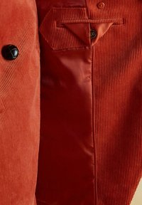 Rūsganas korduroja jaka ar melnu pogu un gludu iekšējo oderi, rādot detalizētu šuvju un auduma tekstūru.