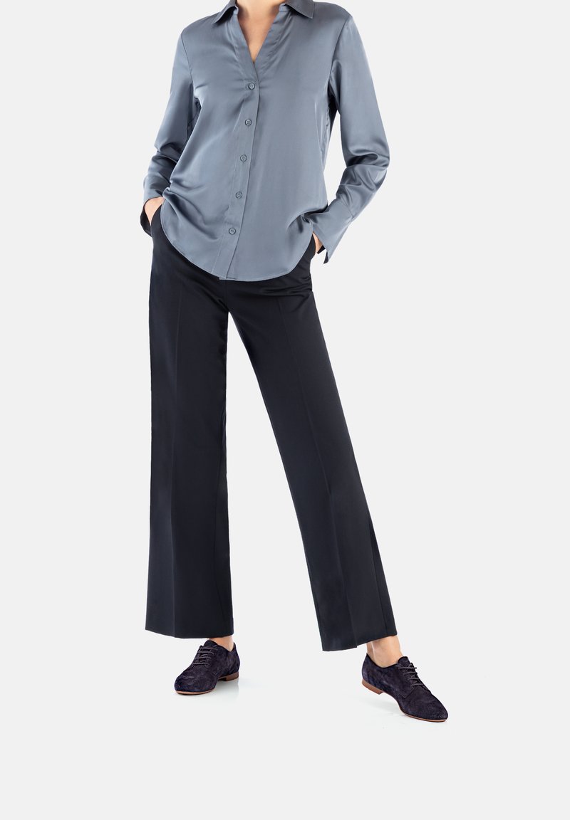 Camicia button-up in satin grigio chiaro con scollo a V; abbinata a pantaloni larghi neri e scarpe stringate in camoscio scuro.