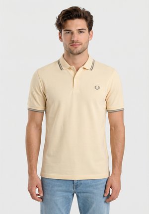 Giovane uomo con capelli castani corti che indossa una polo beige con colletto e polsini a righe, abbinata a jeans azzurri, in piedi.