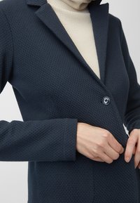 Blazer texturizado azul marino con cuello con muesca, cierre de un botón y mangas largas, superpuesto a un suéter de cuello alto color crema.