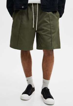 Homme portant un short à cordon de serrage vert olive, des chaussettes blanches, des baskets basses noires et une veste noire partiellement visible sur un fond blanc.