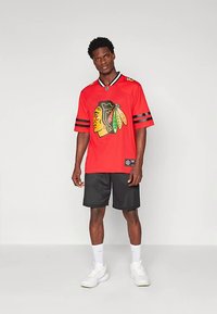 Sarkans krekls ar melnām svītrām, uz tā krāsaina logotipa attēlojums, valkāts kopā ar melnām acīs tīklošanas šortiem. Modelis stāv uz neitrālas fona.