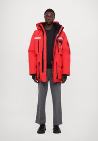 Rote isolierte Parka mit reflektierenden Akzenten, mehreren Taschen, schwarzem Reißverschluss und Besatz, kombiniert mit grauen Hosen und schwarzen Schuhen.