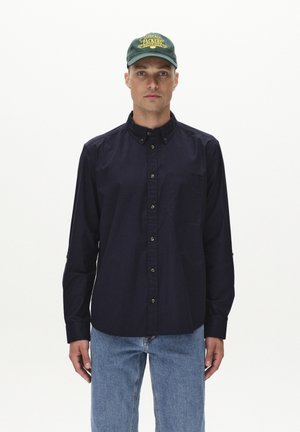 Navy button-up shirt with a pocket, featuring a textured fabric and brown buttons. Paired with light blue denim jeans and a green cap.