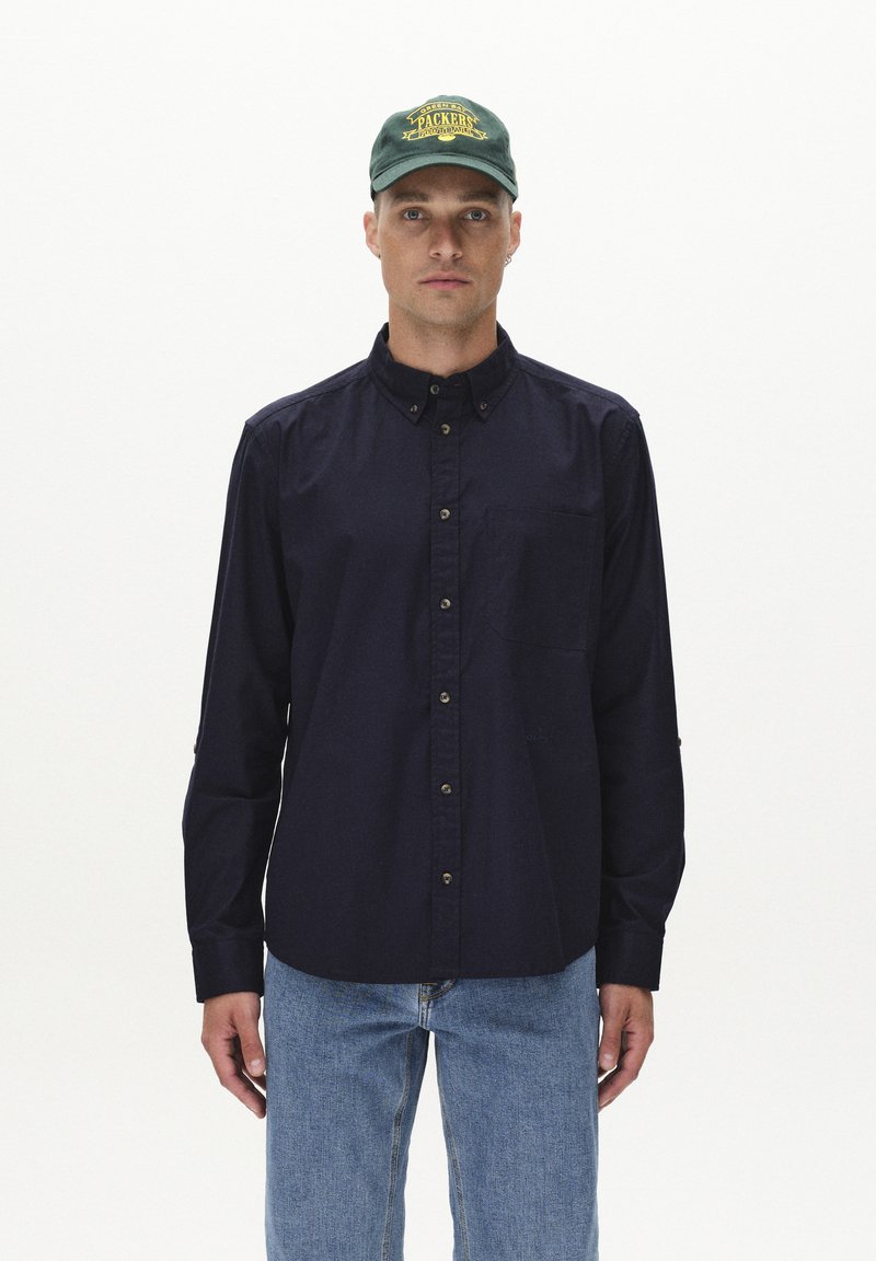 Camicia navy con bottoni, dotata di tasca, realizzata in un tessuto texture e con bottoni marroni. Abbinata a jeans denim azzurri e un cappellino verde.