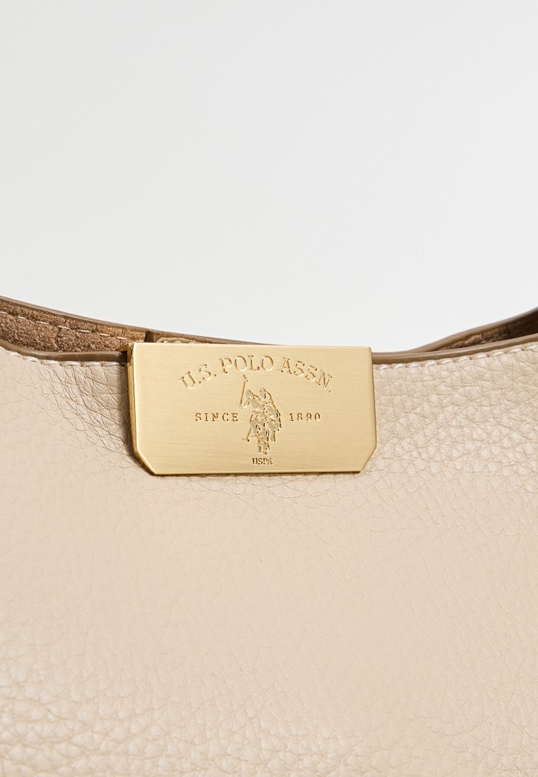 Sezione di una borsa in pelle beige a trama con un'etichetta rettangolare in metallo dorato incisa "U.S. Polo Assn. Dal 1890" e un'illustrazione del logo.
