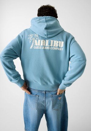 Person, die einen hellblauen Hoodie mit der Aufschrift „Malibu Guess Jeans Company“ und einem Palmen-Motiv auf dem Rücken trägt, kombiniert mit blauen Jeans und von hinten zu sehen ist.