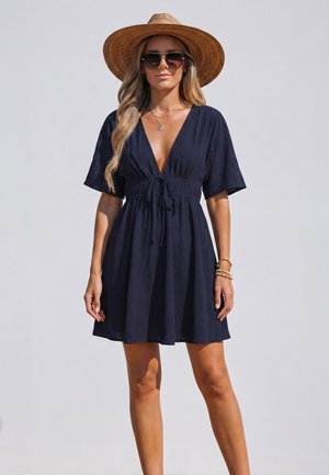 Frau trägt ein marineblaues kurzes Kleid mit tiefem V-Ausschnitt, breitkrempigen Strohhut, Sonnenbrille und Goldschmuck vor einem schlichten hellen Hintergrund.