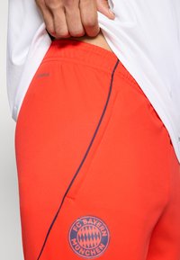 Pantalones cortos deportivos rojos con una costura lateral acentuada en azul y un logo redondo del FC Bayern München. El material parece suave con una cintura elástica.