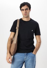 Camiseta de algodón negra con un bolsillo en el pecho que presenta un logotipo blanco. Combinada con unos pantalones vaqueros de denim azul claro y un bolso de hombro de ante marrón.