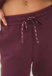 Bordeaux sweatpants med en blød tekstur, elastisk talje og snoretræk med et logo. Tæt på vinkel, der viser detaljerne på taljen.