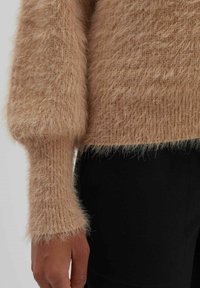 Gros plan d'une personne portant un pull beige duveteux à manches longues et un pantalon noir, mettant en valeur le tissu doux et texturé au niveau du bras et du torse.