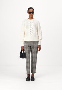 Fleckiger, cremefarbener Strickpullover mit Kabelmuster, hoch taillierte graue Karohosen, schwarze Tasche und Loafers; das Modell trägt schwarze Sonnenbrillen.