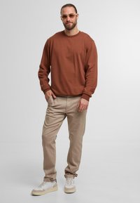 Brown sweatshirt with ribbed cuffs, beige trousers, and white sneakers with light accents; front view of a standing male model.