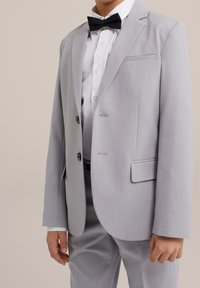 Ljusgrå kostymjacka med två knappar, enkel bröstficka och slanka slag, ihopparad med vit skjorta och svart fluga. Måttlig textur.