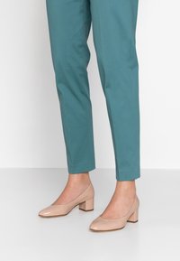 Beige lackskinnsblockklackskor med rund tå, parat med tealfärgade byxor till ankeln. Klacken har en måttlig höjd.