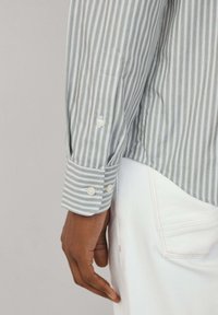 Camicia a righe in misto cotone verde e bianco; presenta polsi con bottoni bianchi, dettagli ai polsi e una morbida texture.