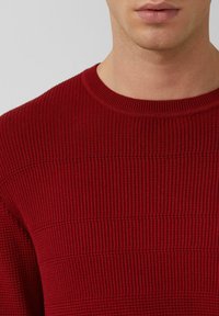 Roter gestrickter Pullover mit horizontaler Rippstruktur und klassischem Rundhalsausschnitt, der im Halsbereich teilweise sichtbar ist.