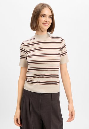 Jonge vrouw met kort lichtbruin haar draagt een beige gestreept T-shirt met korte mouwen en donkere hoge taille broek, glimlacht licht, tegen een witte achtergrond.