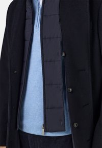 Tumesinise villane mantel helesinise kudumi peal. Mantlil on suured nööbid ja nööbitud, tikitud sisekiht. Näha on tekstuurse kanga detail.