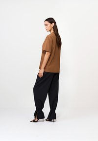Kobieta stojąca w profilu, ubrana w brązowy oversize'owy t-shirt, czarne luźne spodnie z wiązaniami przy kostkach oraz czarne sandały na wysokim obcasie.