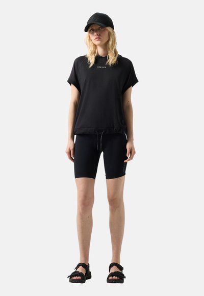 Femme portant une casquette noire, un sweat à capuche noir ample à manches courtes, un cuissard de cyclisme noir et des sandales noires, debout devant un fond clair uni.