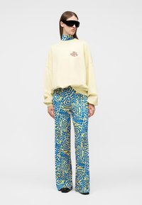 Γυναίκα με υπερμεγέθη κρεμ φούτερ, μπλε και κίτρινες παντελόνες φαρδιάς γραμμής με αφηρημένο μοτίβο, μαύρα γυαλιά ηλίου, στέκεται μπροστά σε λευκό φόντο.