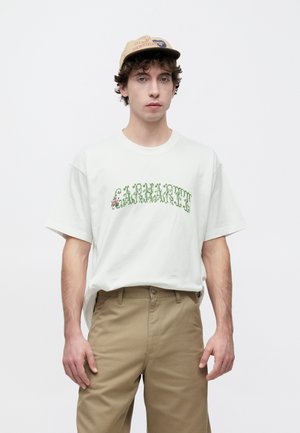 White cotton T-shirt with green and pink graphic text design. Paired with light brown pants, model stands against a neutral background.