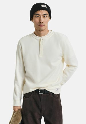 Chemise à manches longues en tricot gaufré crème avec un plastron à boutons et un col rond, associée à un pantalon sombre et un bonnet noir.