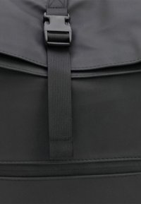 Zwarte rugzak met een bovenklep voorzien van een plastic gesp, nylon schouderband en een gladde synthetische textuur; bevat zichtbare stiksels en een rits.