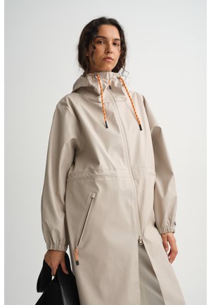 Ženska v bež deževni jakni s kapuco in oranžnimi vrvicami drži črno torbico, stoji pred enobarvnim belim ozadjem.