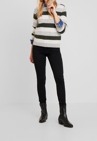 Suéter a rayas en mezcla de lana gris y verde oliva, con mangas de 3/4 sobre una camisa de denim azul, combinado con jeans ajustados negros y botas de tobillo.