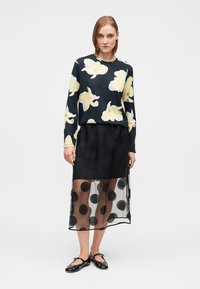 Μαύρη μακριά φούστα με διάφανο πουά επικάλυμμα, συνδυασμένη με μπλε πουλόβερ με κίτρινα λουλουδένια σχέδια. Το μοντέλο φοράει μαύρα επίπεδα παπούτσια.