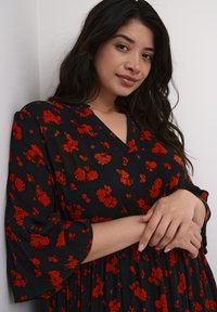 Žena s dlouhými tmavými vlasy v černých šatech s červeným květinovým vzorem, stojící proti bílé zdi s jemně spojenýma rukama.