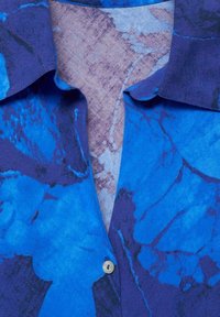 Blaue Hemd mit einem floralen Muster in verschiedenen Blau- und Lilatönen. Mit Kragen und einem hellen Knopf, weicher Textur.