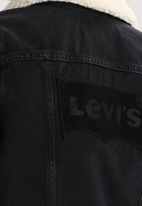 Veste en jean noire avec col en laine. Présente un patch logo Levi's texturé noir au dos. Design lisse et structuré.