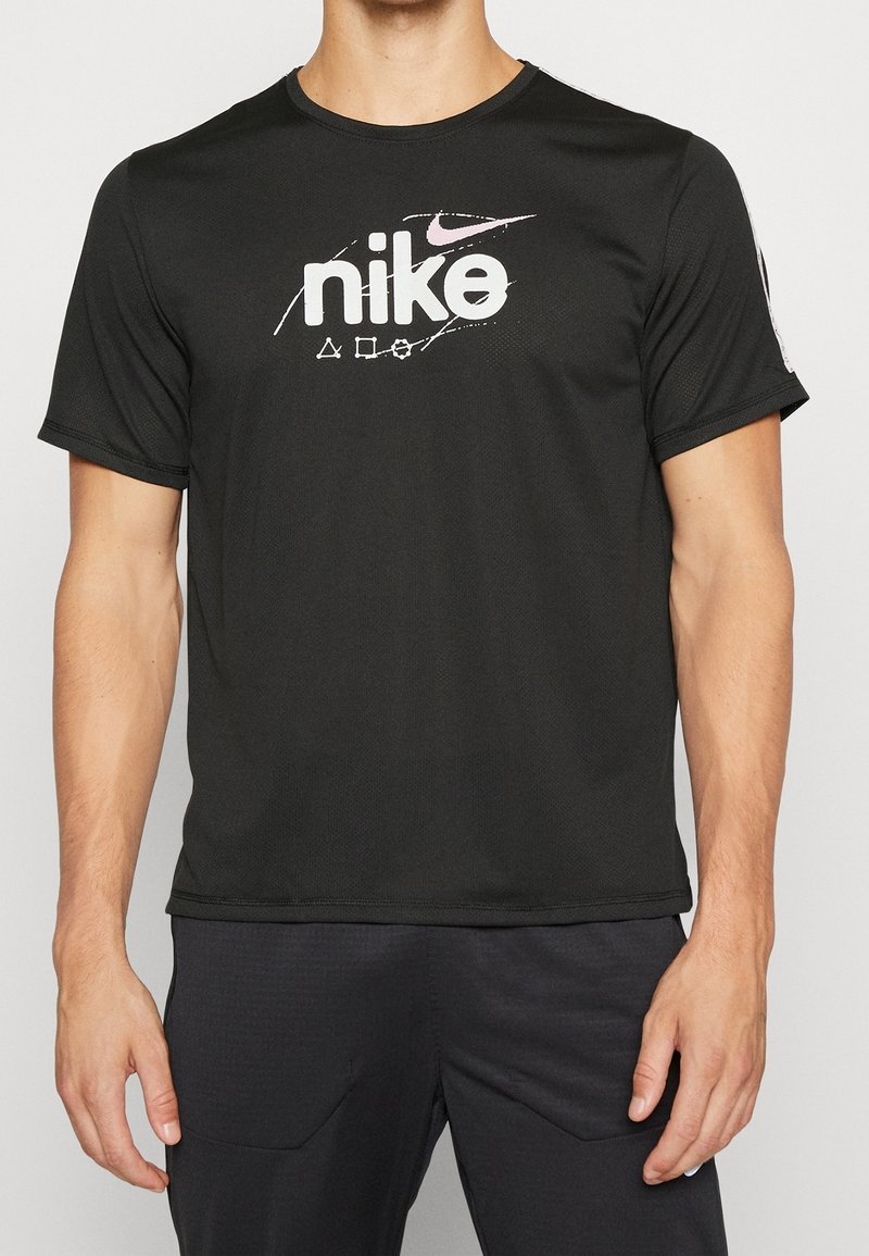 Man med en svart kortärmad Nike-t-shirt med vit och rosa logotyp och symboler på bröstet, kombinerat med svarta byxor.