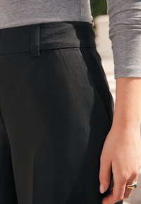 Mustad kohandatud püksid, millel on sile tekstuur ja lamedapoolne vöökoht. Käsi toetub kangale, näidates sõrmel sõrmust.