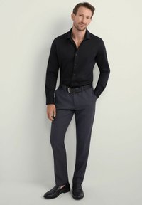 Czarna koszula z długim rękawem i zapięciem na guziki, zestawiona z ciemnoszarymi eleganckimi spodniami. Buty to czarne mokasyny. Neutralne tło.