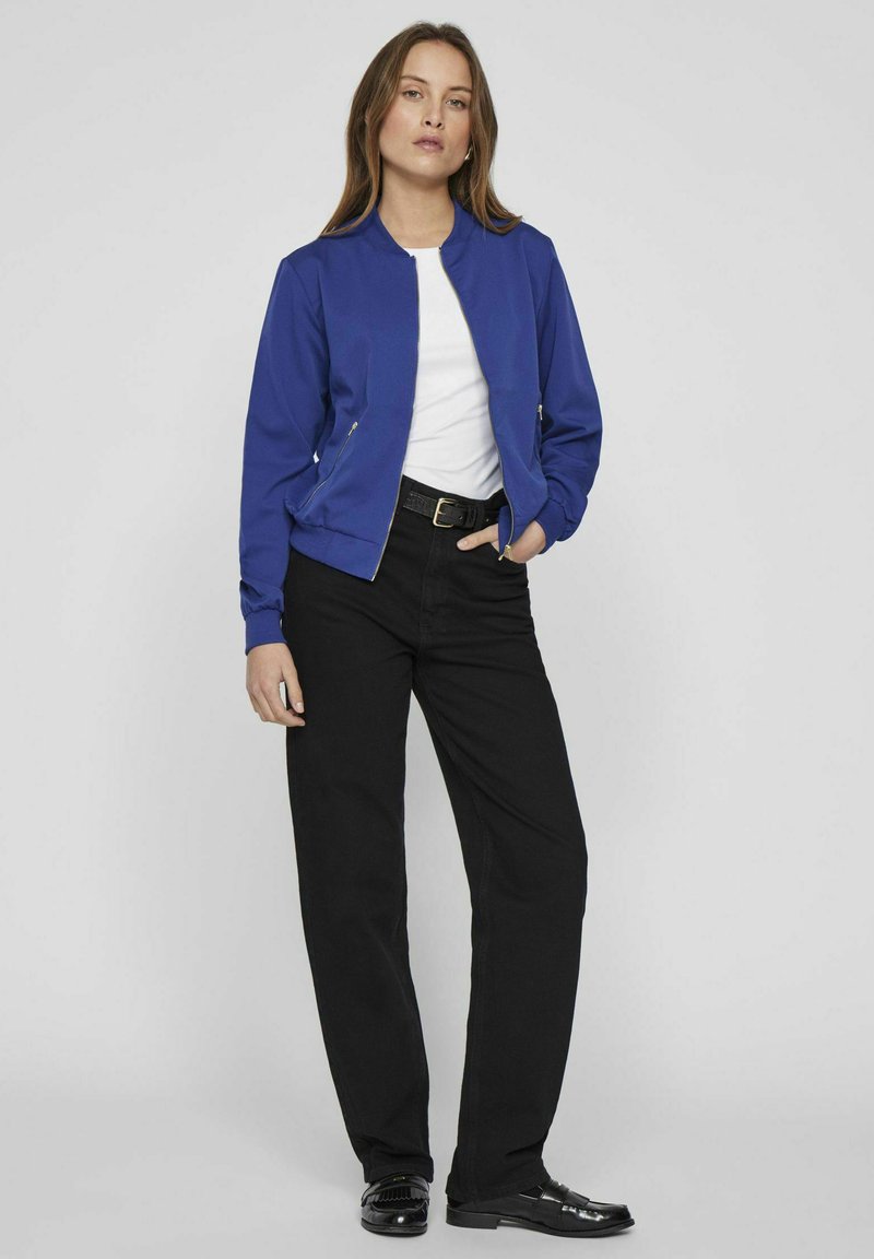 Niebieska bomberka z zamkiem z przodu, ściągaczami przy mankietach oraz kieszeniami, noszona na białej koszuli i zestawiona z czarnymi spodniami prostymi.