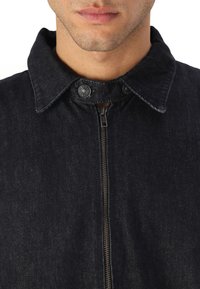 Button-up denim jacket in dark blue. Features a collar, front zipper, and snap details. Fabric has a textured appearance.