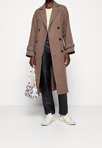 Brun uldfrakke med sorte knapper, parret med sorte læderbukser og hvide sneakers med gule detaljer. Holder en taske med blomsterprint.