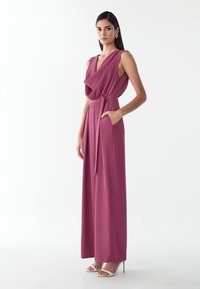 Femme portant une combinaison sans manches mauve avec un décolleté drapé, une ceinture à la taille, des poches latérales et des sandales blanches à talons hauts sur fond blanc.