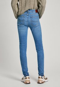 Jeans ajustados de mezclilla azul con talle medio, bolsillos traseros con parche y detalles sutiles de costura, combinados con zapatillas deportivas blancas y doradas.