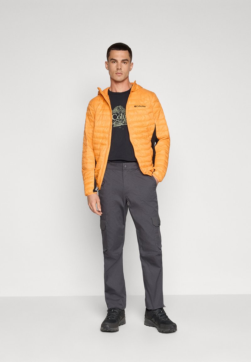 Veste matelassée orange avec une capuche, assortie d'un t-shirt graphique noir et d'un pantalon cargo gris, complétée par des chaussures noires.