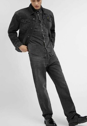 Junger Mann trägt schwarze Denimjacke, schwarzes Hemd mit Knöpfen, dunkle Jeans und schwarze Schuhe, posiert mit einer Hand in der Tasche, weißer Hintergrund.