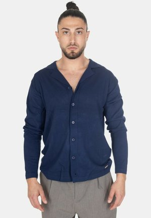 Uomo con capelli scuri raccolti in uno chignon, che indossa un cardigan blu navy con bottoni e pantaloni grigio chiaro, in piedi su uno sfondo uniforme.