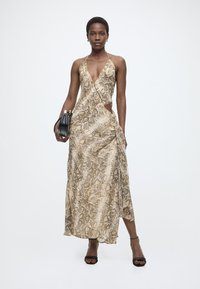 Γυναίκα που περπατά φορώντας αμάνικο, μακρύ φόρεμα με μπεζ σχέδιο φιδιού, κρατώντας μαύρο τσαντάκι χειρός, φορώντας μαύρες σανδάλες στιλέτο και χρυσά σκουλαρίκια.