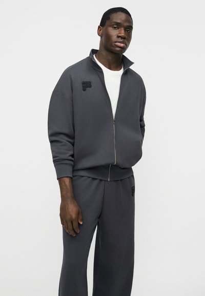 Homme portant une veste zippée gris foncé et un pantalon assorti, debout les mains détendues, sur un fond blanc uni.