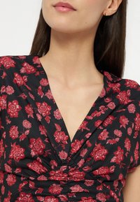 Γυναίκα με μακριά σκούρα μαλλιά που φοράει μαύρο τοπ με κόκκινο λουλουδάτο μοτίβο και στρογγυλεμένο V-λαιμόκοψη, εμφανίζοντας χρυσά σκουλαρίκια καρφιά.